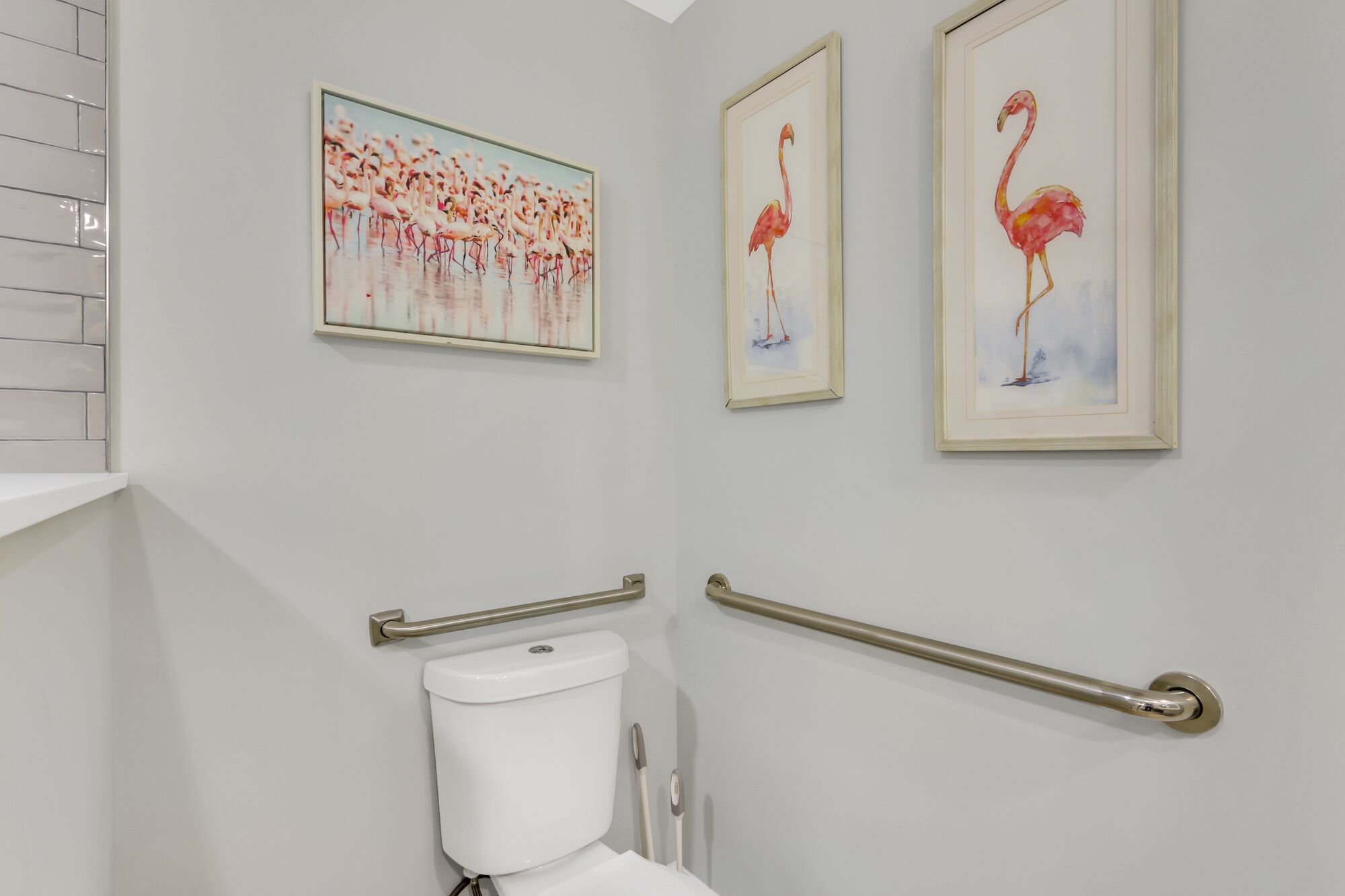 A bathroom with a toilet, a handrail, and three framed paintings of flamingos on the wall.