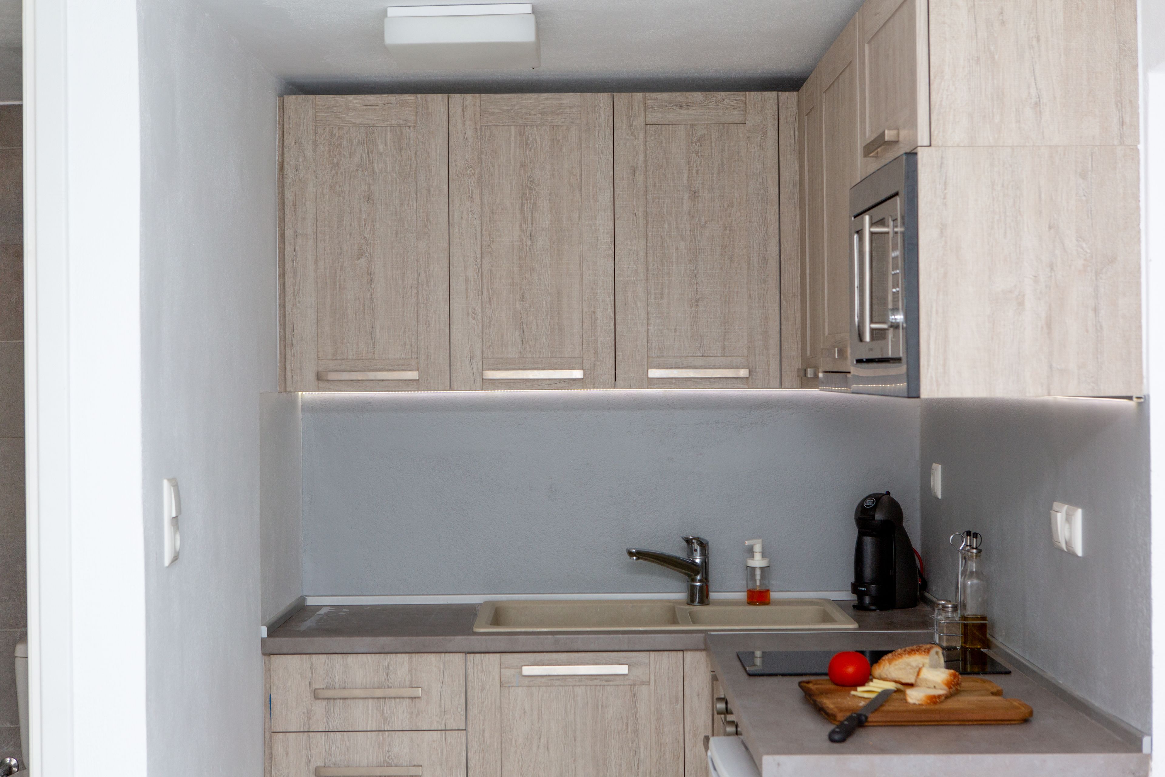 Une cuisine compacte avec des meubles en bois, un évier en acier inoxydable, une machine à café et une planche à découper avec une tomate et un couteau.