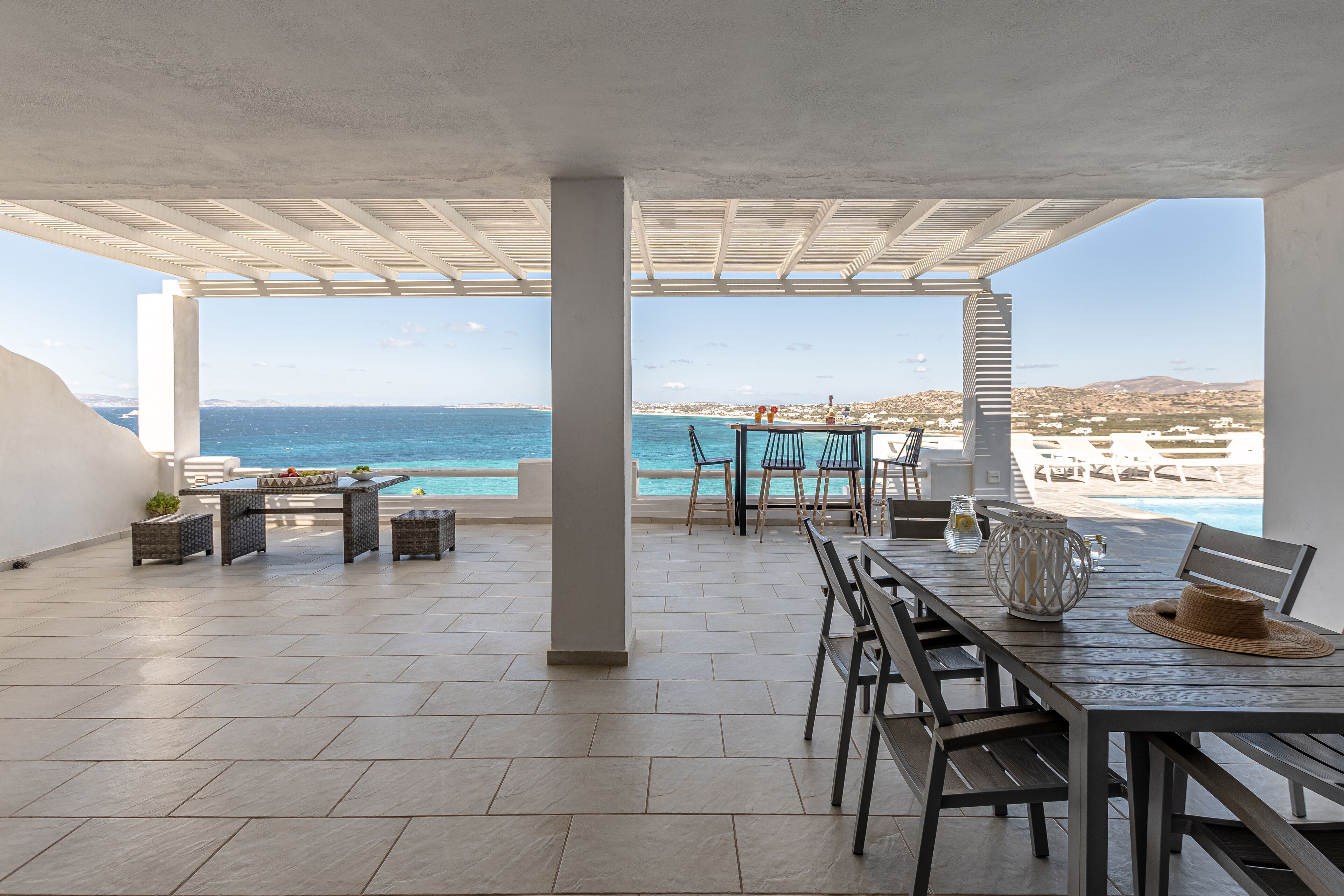 Un espace extérieur abrité, avec vue sur la mer, comprenant une table et des chaises, un vase décoratif et un chapeau posé sur la table.