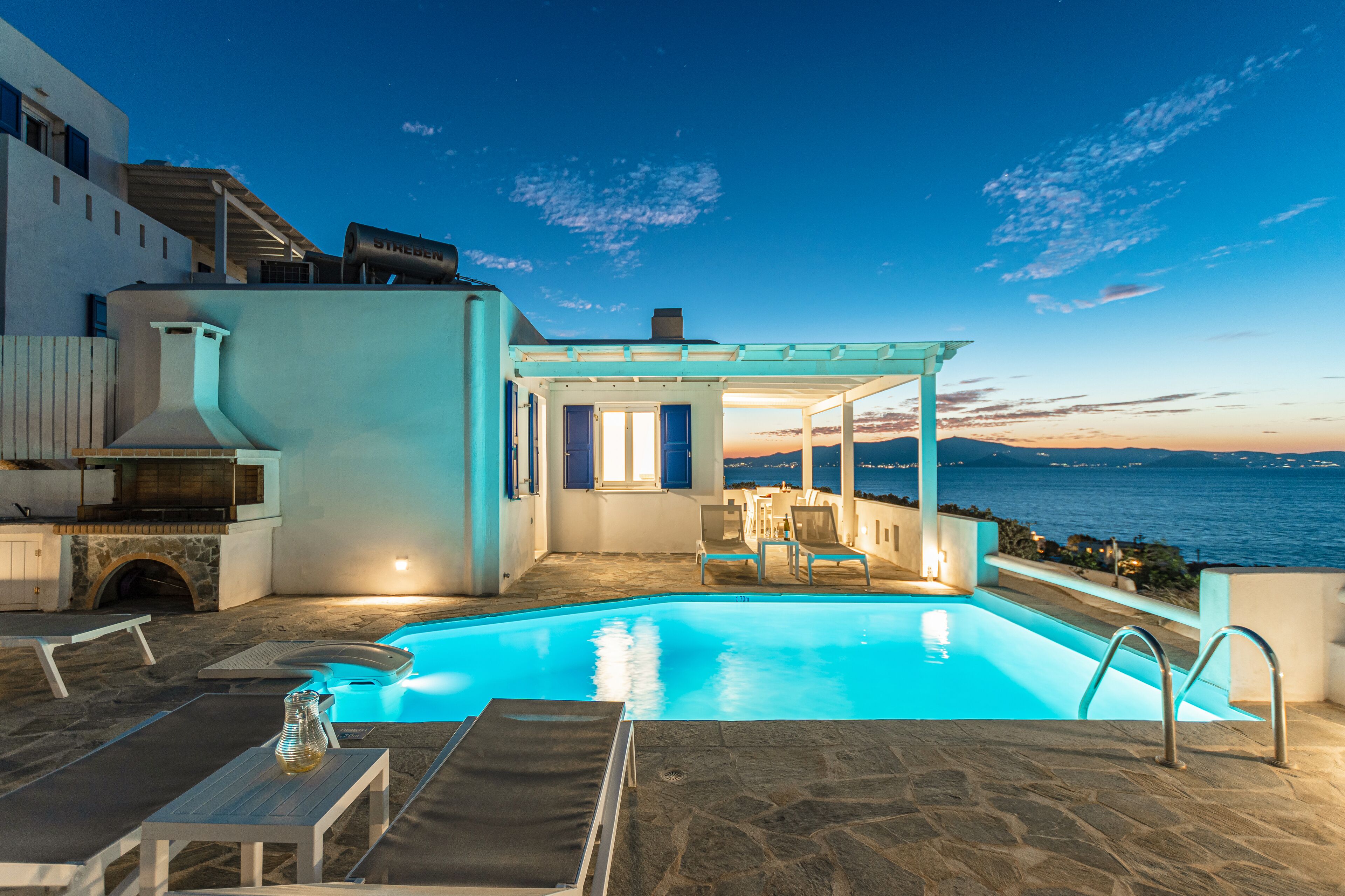 Un espace piscine aménagé avec des chaises longues, une pergola et une vue sur la mer au crépuscule.