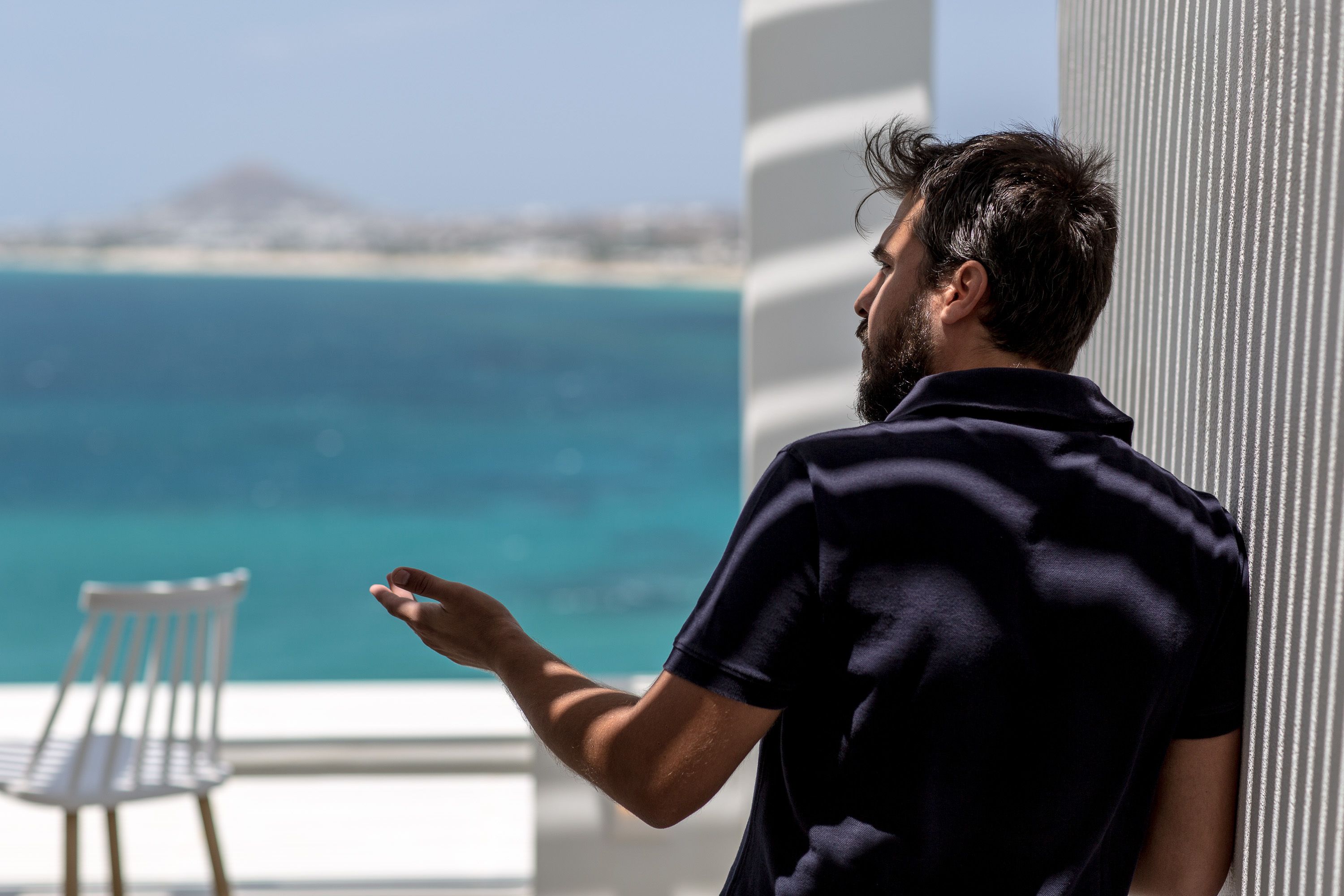 A person standing by a window overlooking a beach with a chair visible inside.