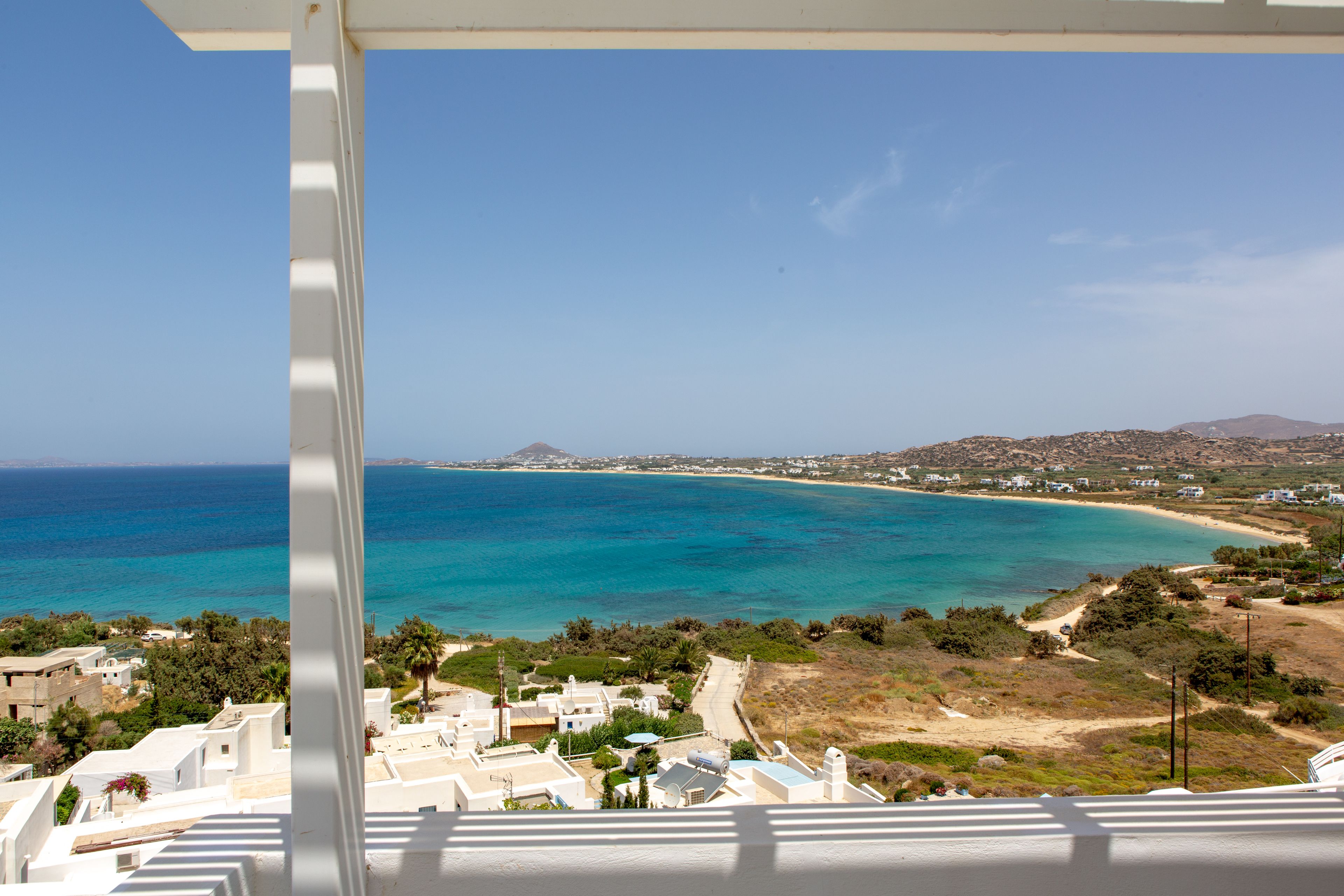 A coastal view from a balcony with a clear blue sea, sandy beach, and distant hills.