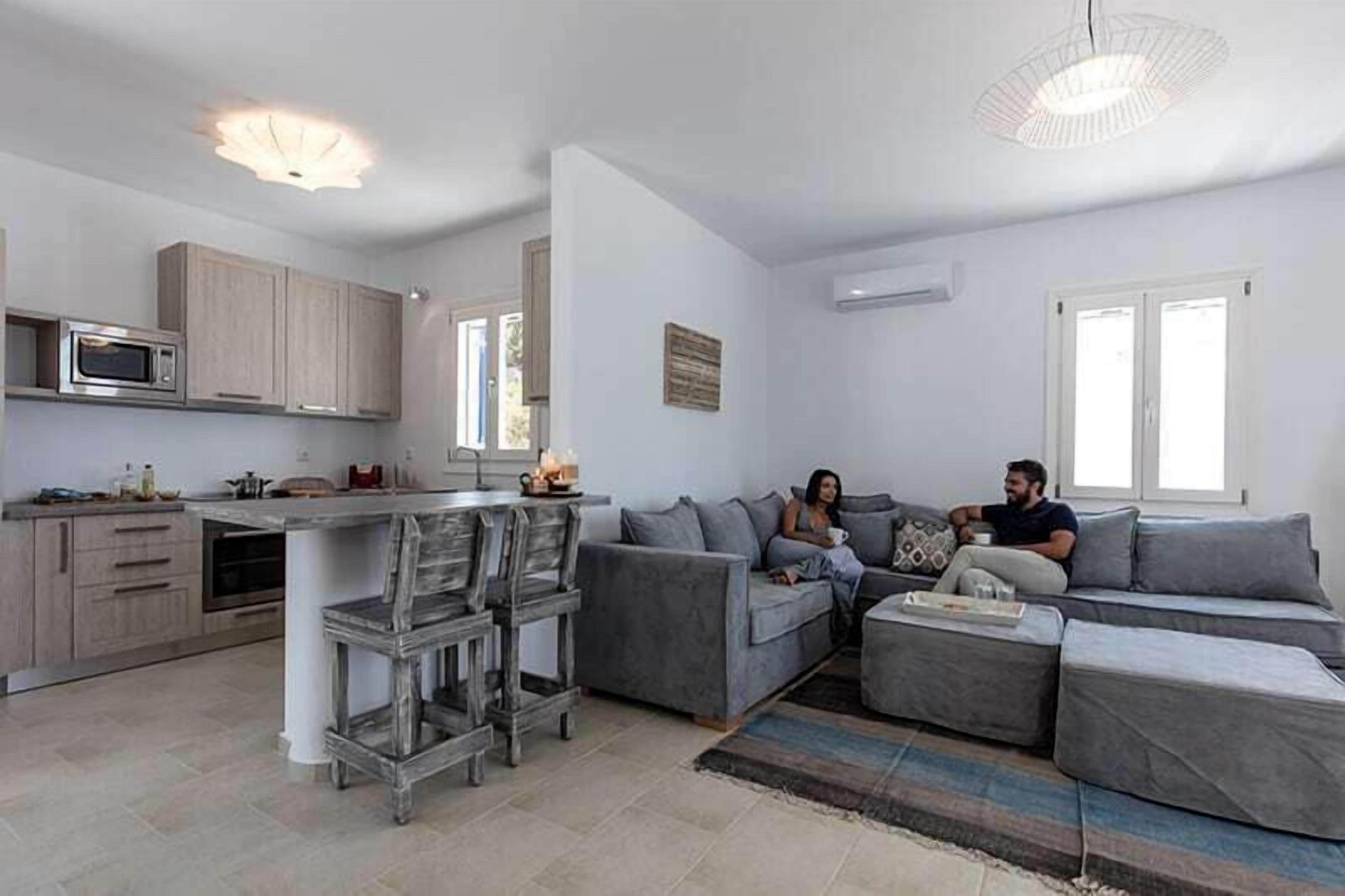 Un soggiorno moderno con un divano grigio, una zona pranzo con un tavolo in legno e una cucina con mobili chiari in legno.