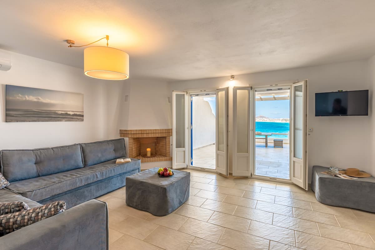 Een moderne woonkamer met een bank, poef en een open haard. Een televisie is aan de muur bevestigd en er is uitzicht op zee door openslaande deuren.