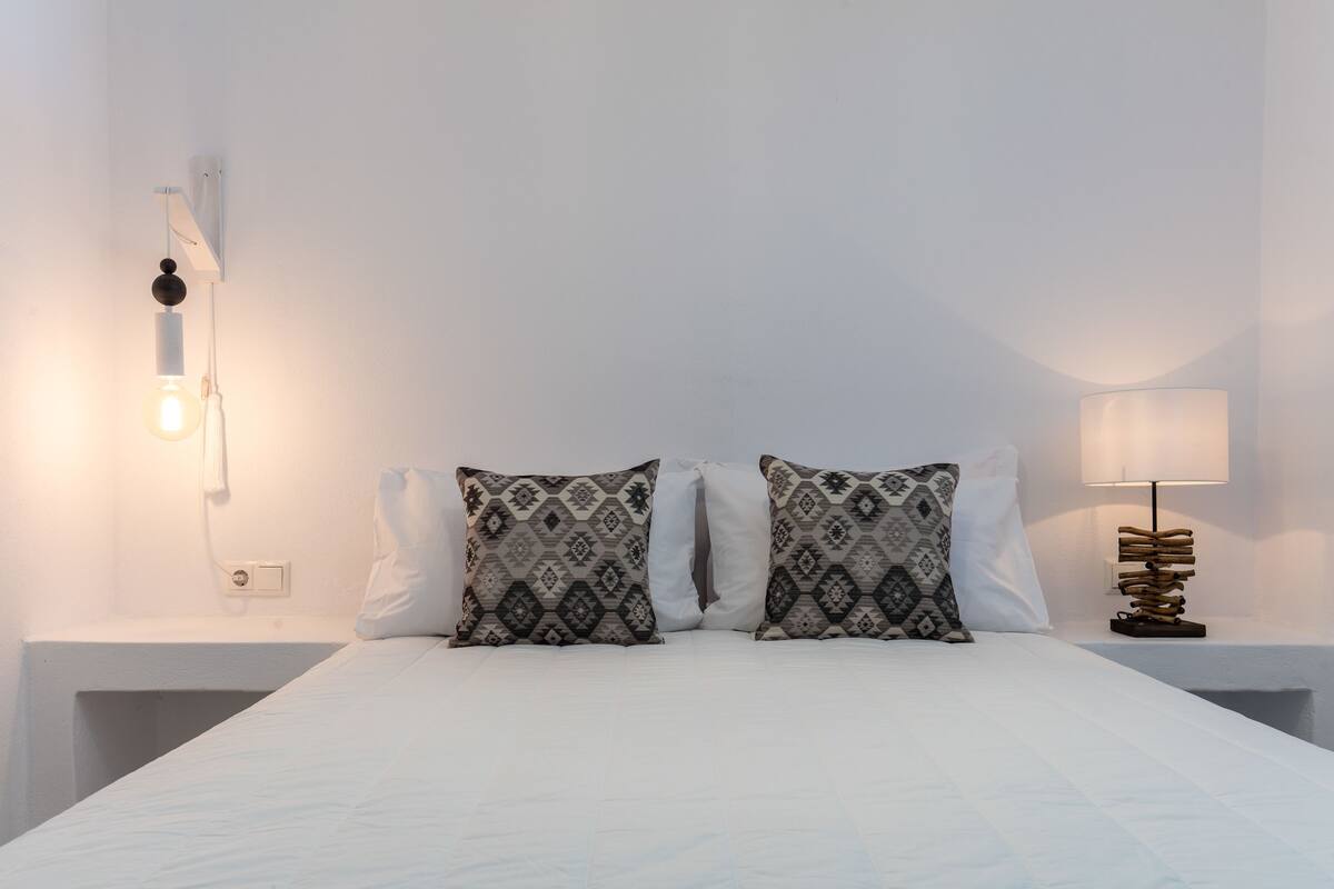 Een net opgemaakt bed met gestreepte kussens, een bedlampje en een wandlamp.