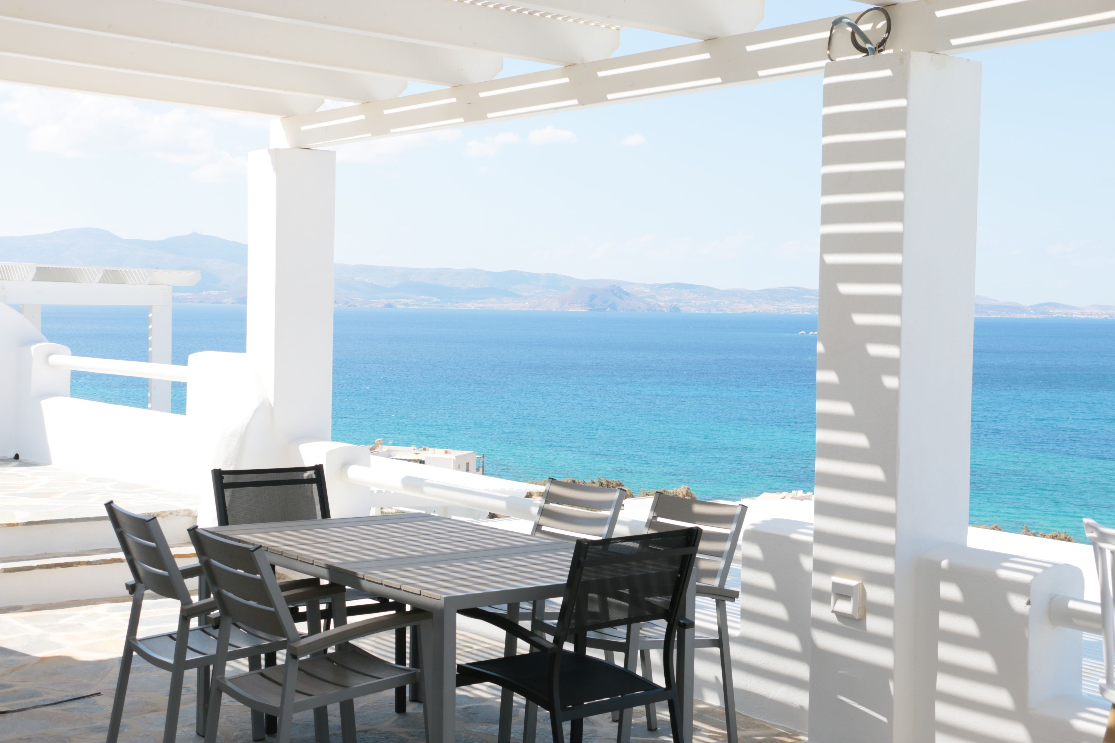 A white outdoor seating area with a table and chairs, overlooking a clear blue sea and distant mountains.
