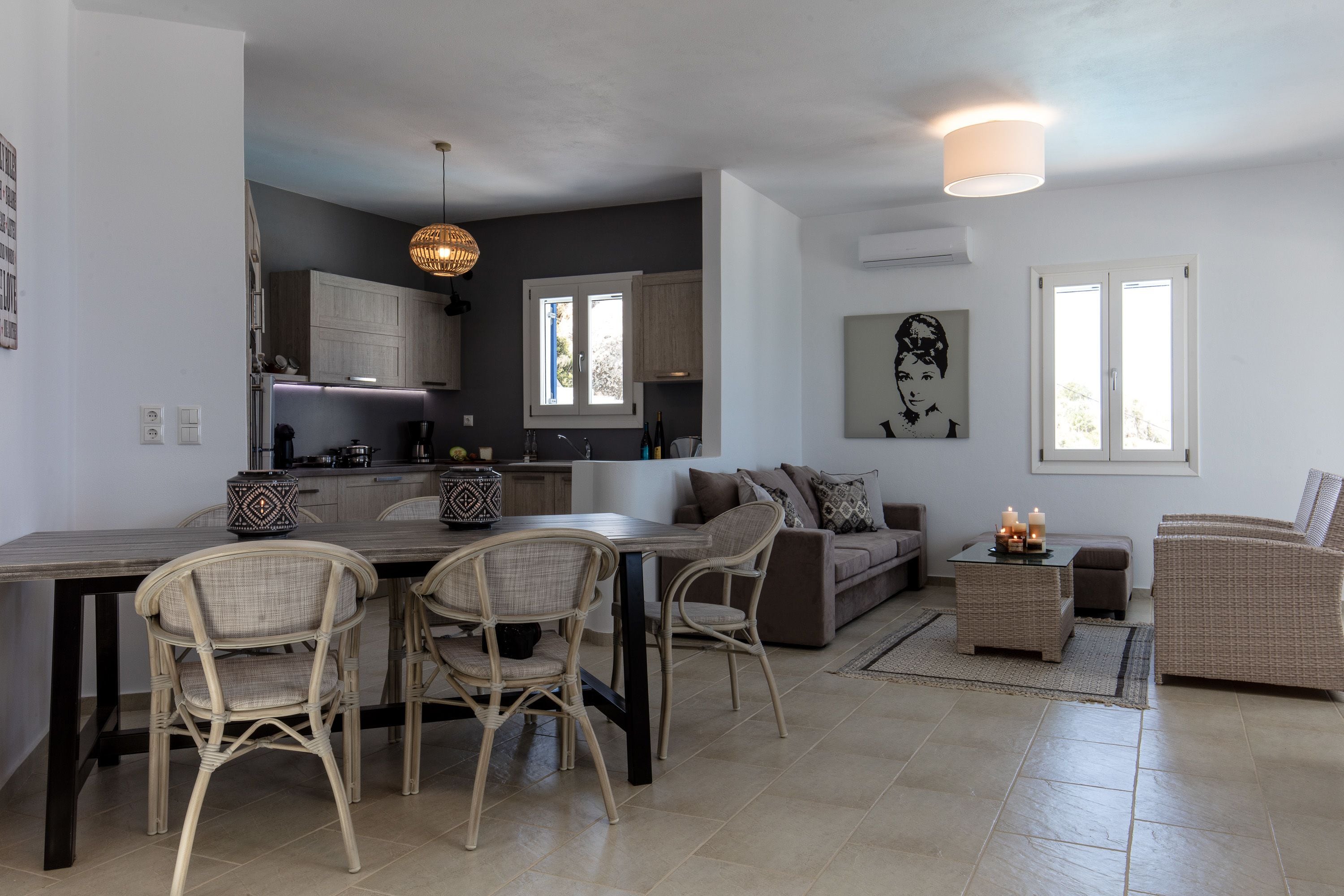 A modern kitchen with dark cabinetry, a dining area with wicker chairs, a sofa, and a small side table with candles.