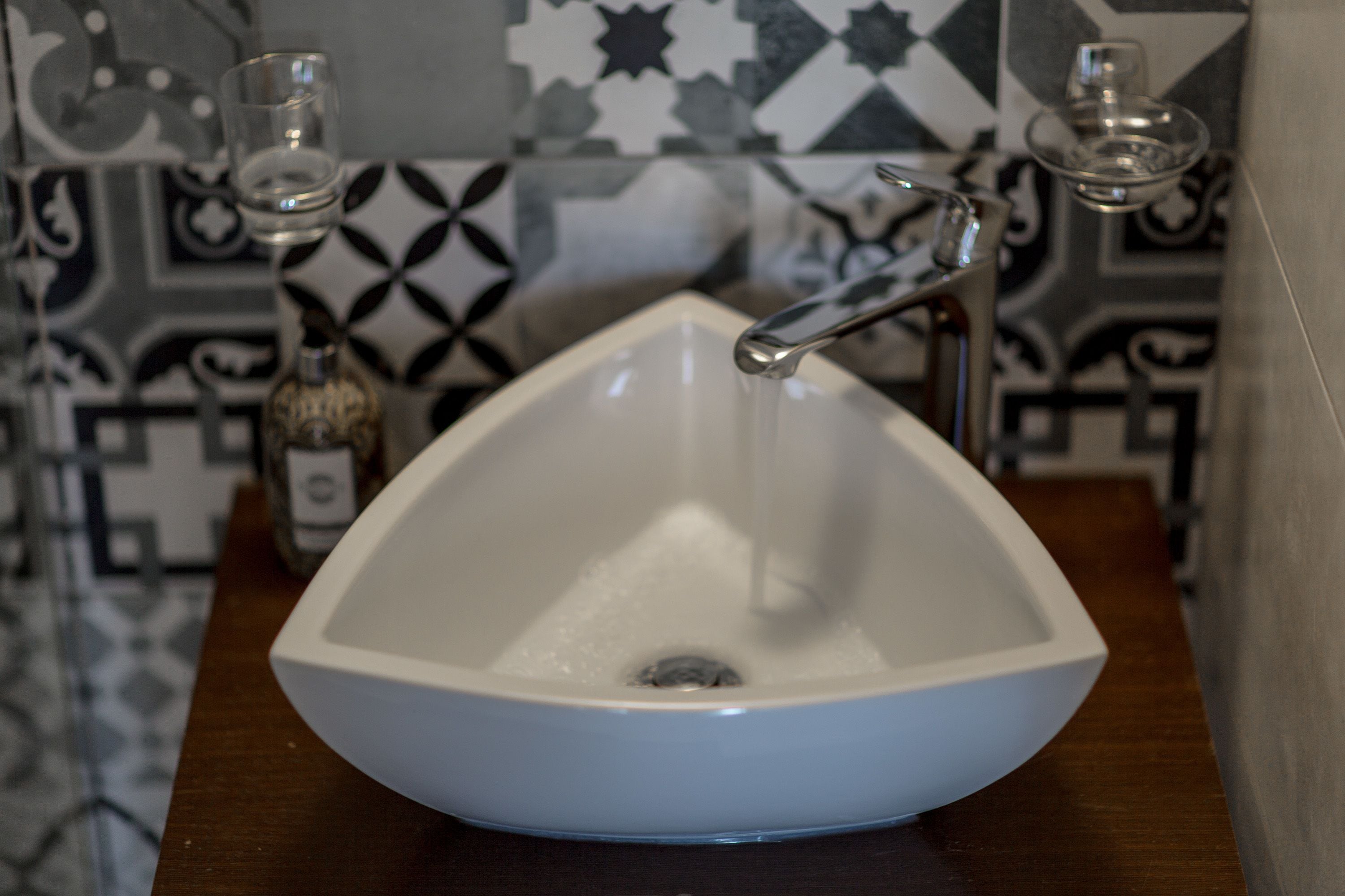 A white, bowl-shaped sink with a single faucet, set against a tiled backsplash with a geometric pattern.