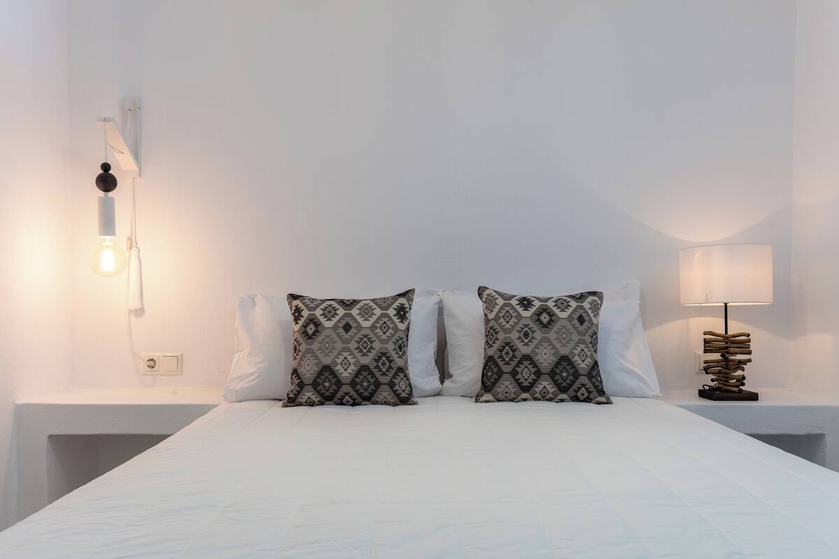 Een net opgemaakt bed met gestreepte kussens, een bedlampje en een wandlamp.