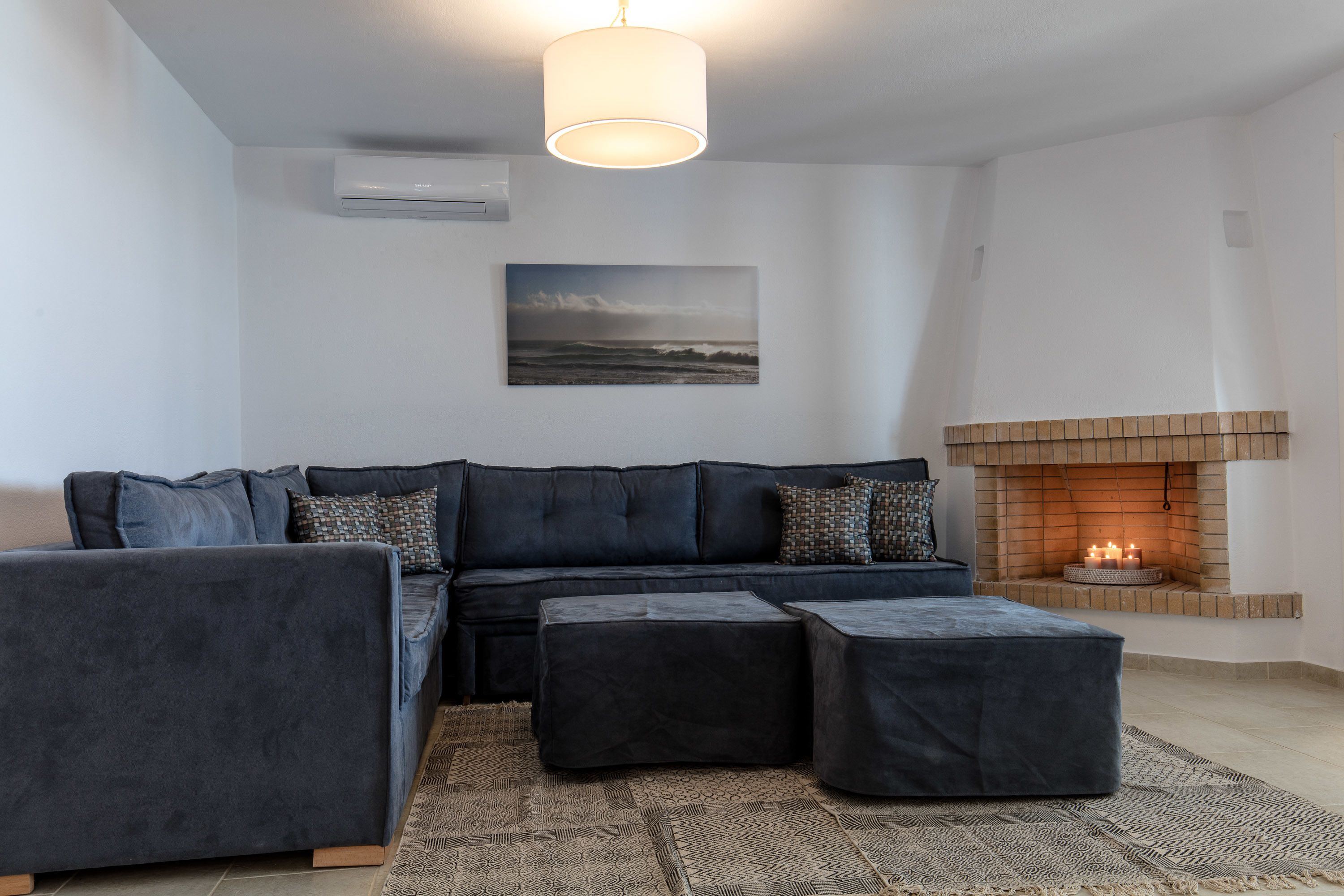 A modern living room with a dark blue sectional sofa, two ottomans, a patterned rug, a fireplace, and a framed landscape painting on the wall.