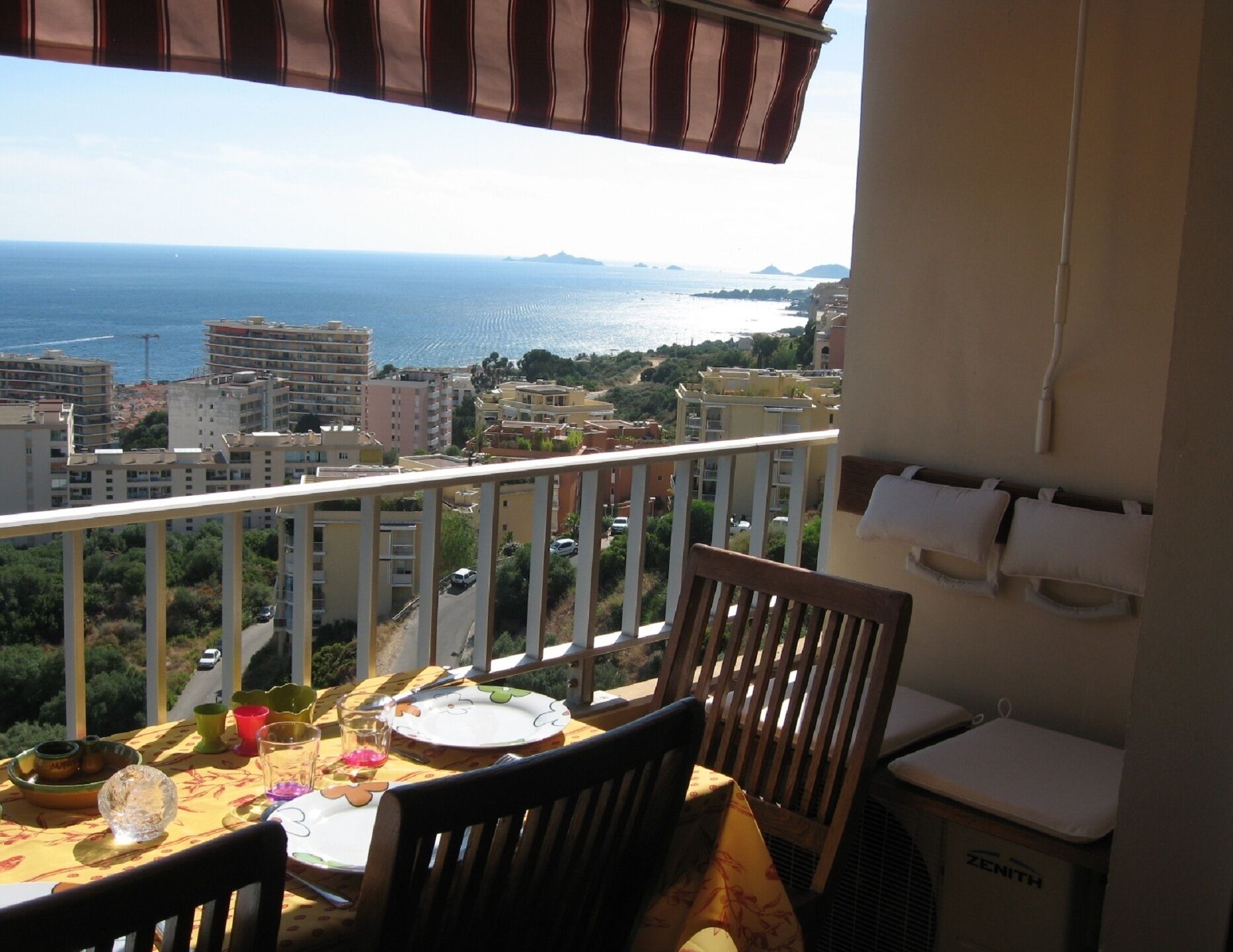 Un balcón con mesa lista para comer, con vistas a un paisaje urbano costero que incluye edificios y el mar.