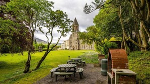 Ein Innenhof mit Picknicktischen und einer Grillgelegenheit aus Backstein, umgeben von üppiger Vegetation und einem historischen Gebäude mit einem Turm im Hintergrund.