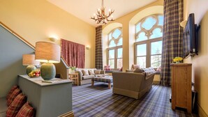 Ein gemütliches Wohnzimmer mit Sofa, Sesseln, einem Couchtisch und einem Fernseher. Eine Treppe befindet sich links, ein Fenster mit Vorhängen sowie eine Deckenleuchte.