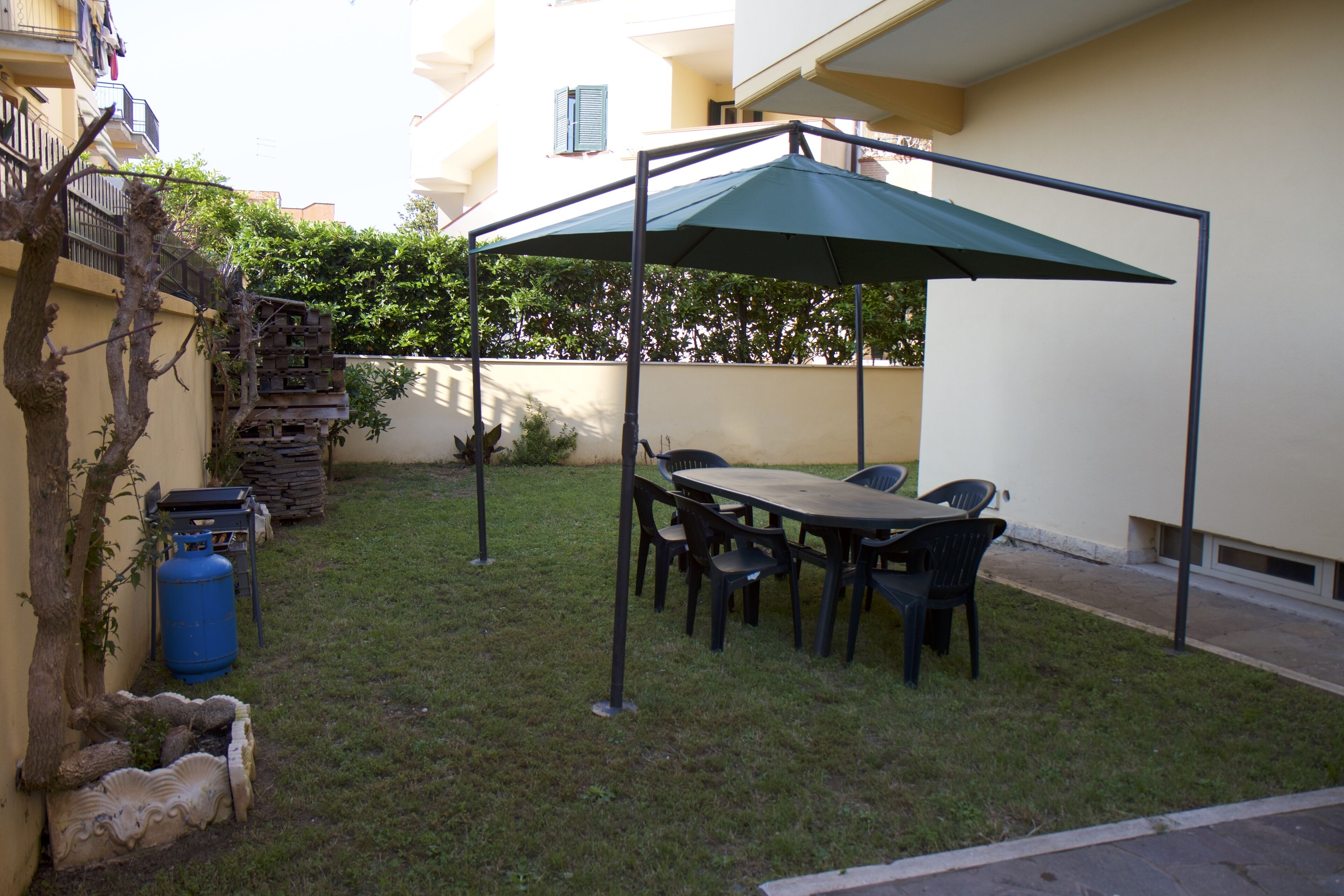 A patio with a table and chairs under a canopy, surrounded by a small garden and a building.