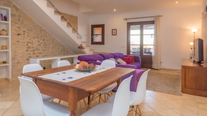 A dining area with a wooden table and white chairs, a staircase with a stone wall, a purple sofa, and a television on a wooden cabinet.
