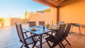 An outdoor patio with a wooden table set for dining, surrounded by chairs, and a grill in the background.