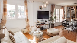 A spacious living room with a large sofa, a glass coffee table, and a flat-screen TV. There is a dining area with a glass table and chairs, and a bookshelf filled with books. - villa near cannes (Mougins)
