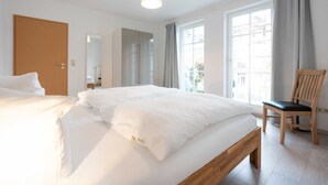 A hotel room with a large bed, a wooden bench, a chair, and a window with curtains.