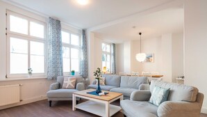 A modern living room with a light-colored sofa, a wooden coffee table, and a large window with patterned curtains. - Starfish - villa on the beach (Heringsdorf)