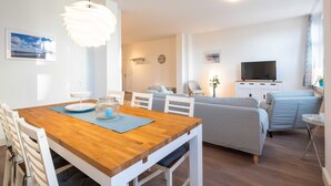 A modern living room with a wooden dining table, white chairs, a sofa, a TV, and a decorative hanging light. - Starfish - villa on the beach (Heringsdorf)