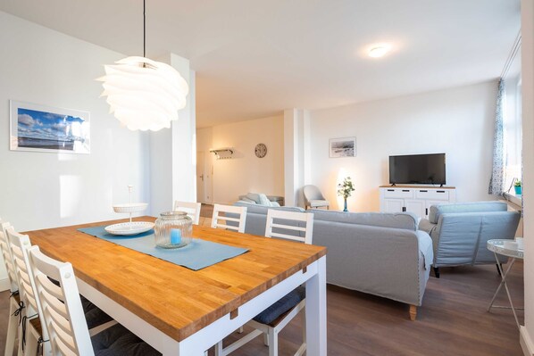 A modern living room with a wooden dining table, white chairs, a sofa, a TV, and a decorative hanging light.