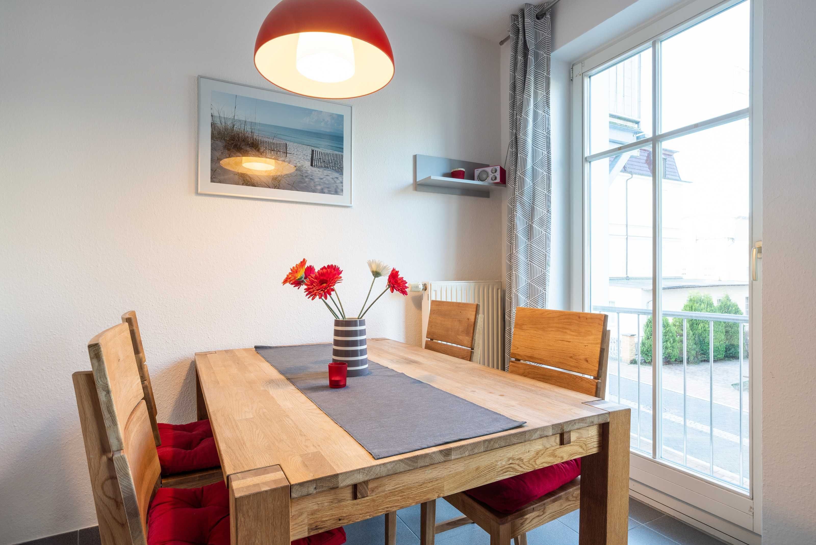 A dining area with a wooden table, red cushions, a vase with flowers, a red lamp, a framed picture on the wall, and a window with a view of the street.