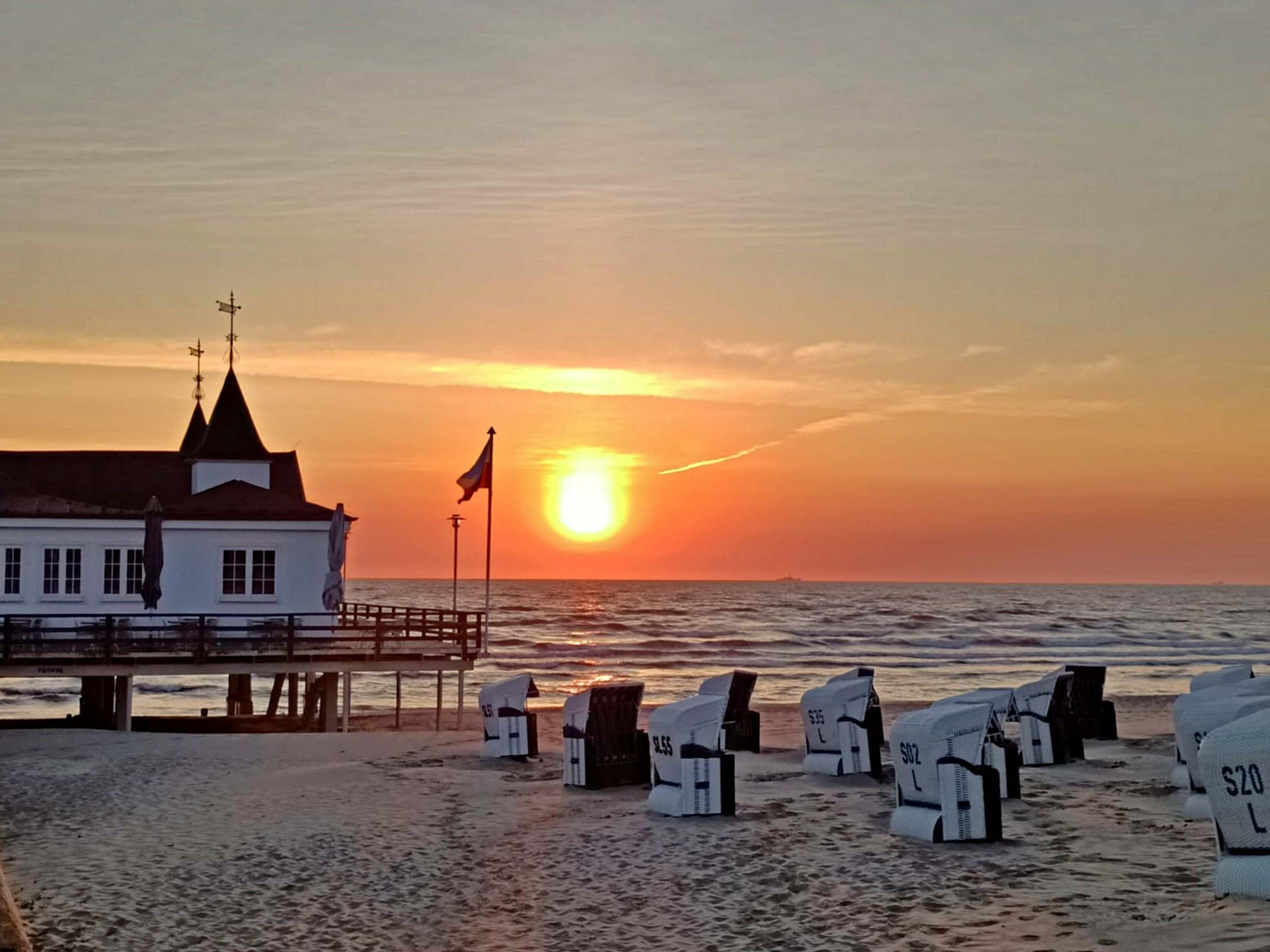 En bygning ved stranden med en mole, strandstole og en solnedgang.