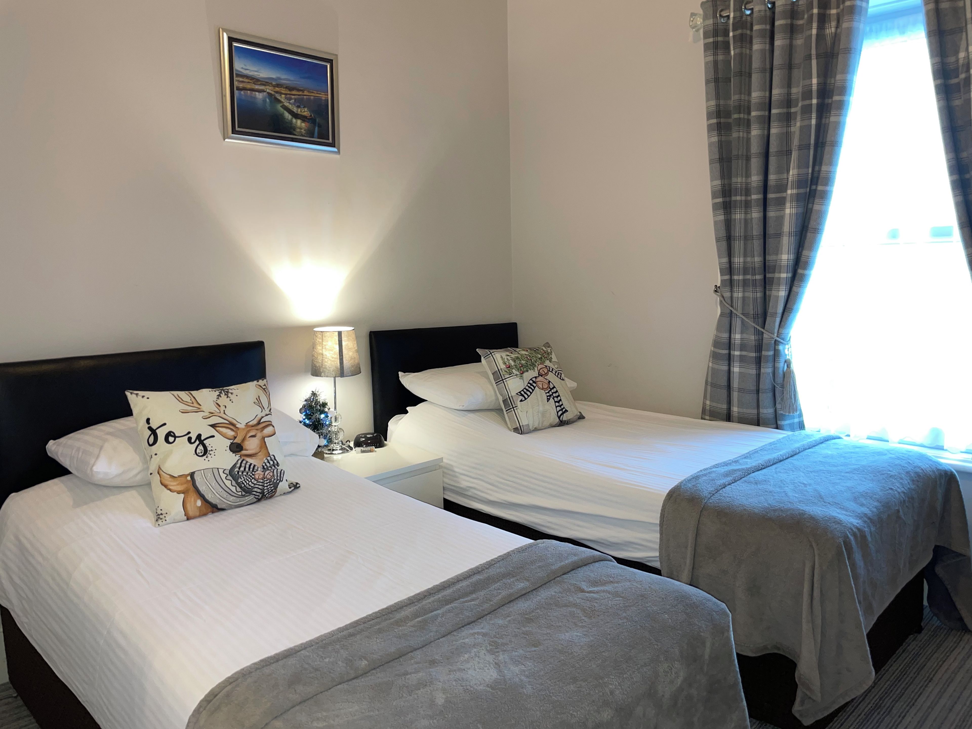 A hotel room with two beds, a nightstand with a lamp, a small Christmas tree, and a framed picture on the wall.