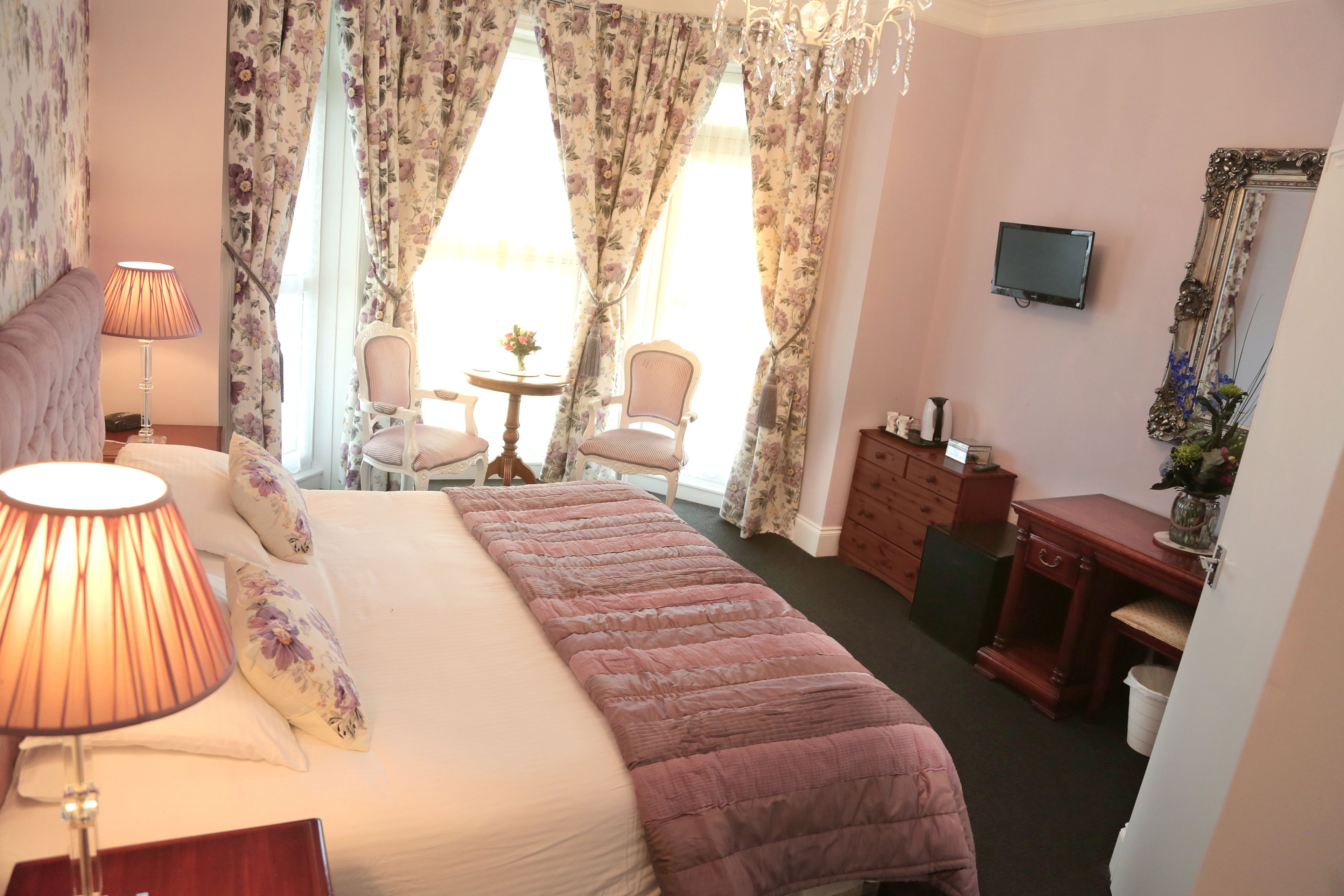A hotel room with a large bed, a bedside lamp, a TV, a dresser, and a chair near the window.