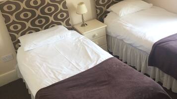 A hotel room with two single beds, each with a patterned headboard, a bedside table with a lamp, and a brown throw blanket on one bed.