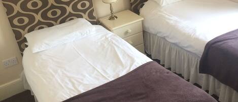 A hotel room with two single beds, each with a patterned headboard, a bedside table with a lamp, and a brown throw blanket on one bed.