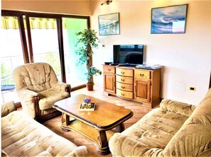 Ein Wohnzimmer mit einer Couch, einem Sessel, einem Holztisch, einem Fernsehmöbel und einem Blumentopf.