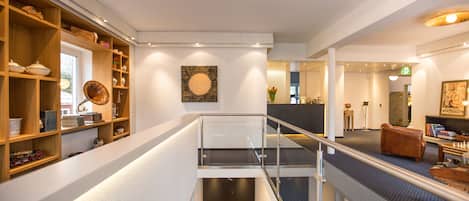 Un interior moderno con una escalera mecánica central, estanterías de madera con varios objetos, y un cuadro abstracto grande en la pared.