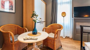 A room with two wicker chairs, a small round table with a vase and a booklet, a wardrobe, a television, and a floor lamp.