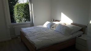 A bedroom with a wooden bed, white bedding, a nightstand, a bedside table, and a window with a view of greenery.
