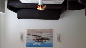A living room with a black leather sofa, a white pendant light, and a painting of a pier.