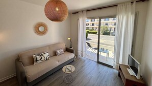 A modern living room with a sofa, a round table, a TV stand, a large window with curtains, and a hanging light fixture. - Cozy 2-Room Apartment, 250m from Beach, Terrace, Private Parking, Sleeps 4 (Port-la-Nouvelle)