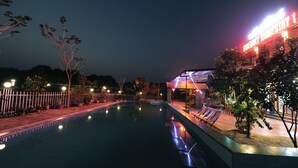 Un área de piscina nocturna con una terraza bien iluminada, sillones de descanso y un edificio con señalización luminosa.