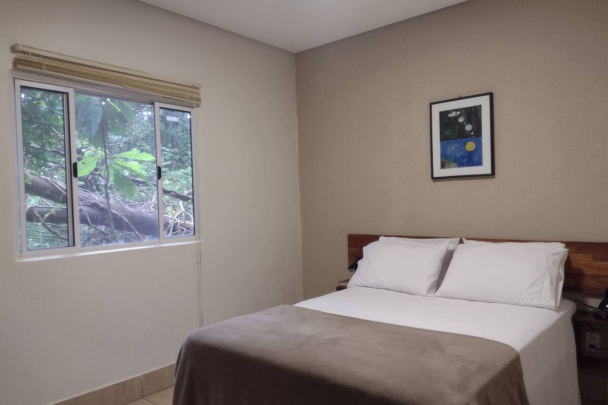 A bedroom with a bed, a window with a view of trees, a bedside table, and a framed picture on the wall.