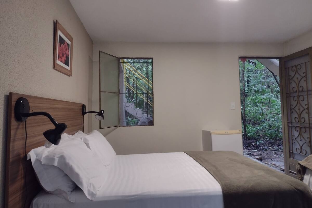 A bedroom with a bed, a bedside lamp, a window with a view of greenery, and a door leading outside.