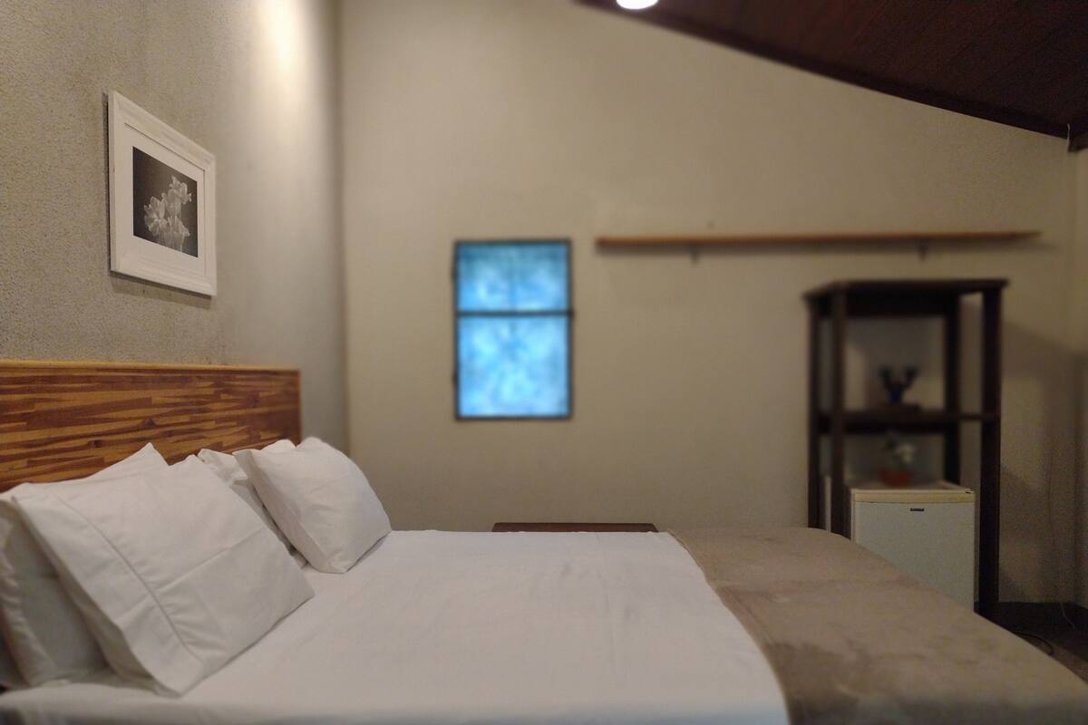 A neatly made bed with white linens in a room with a wooden headboard, a small shelf unit, and a framed picture on the wall.