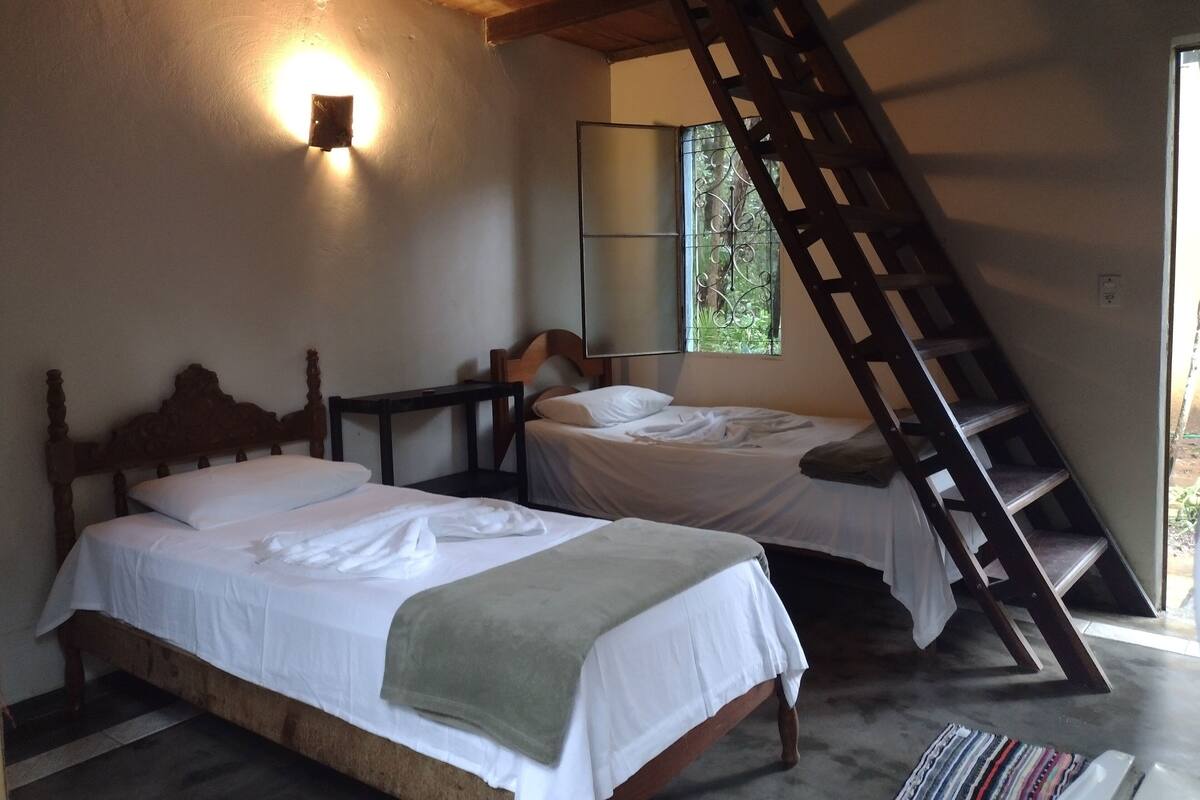 A bedroom with two beds, a wooden ladder, a small table, and a window with a view of greenery.