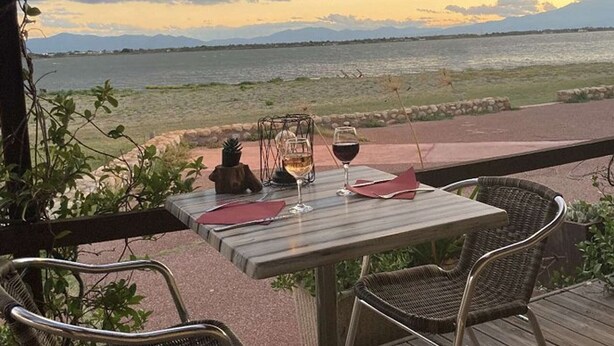A table with two glasses of wine, a candle, and a menu on a terrace overlooking a body of water and mountains. - Elevated Ground Floor Apartment, 3 Rooms, 6 Sleeps, Pool, Terrace, Parking, Sports Facilities (Port Barcarès)