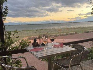 Une table avec deux verres de vin, une bougie et un menu, sur une terrasse donnant sur un plan d’eau et des montagnes.