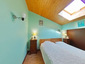 Un dormitorio con techo de madera, tragaluz, cama con colcha estampada, dos mesitas de noche con lámparas y una lámpara de pared.