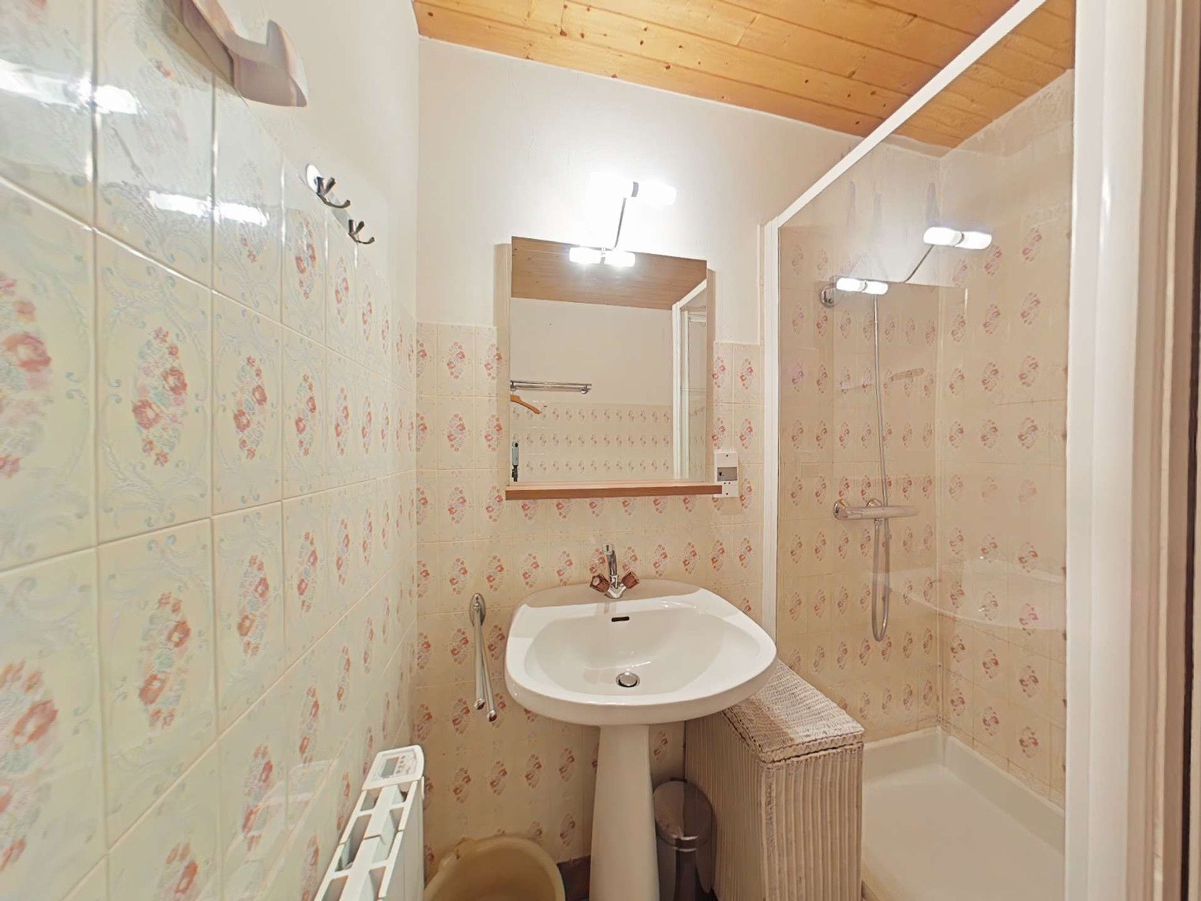 A bathroom with floral patterned tiles, a pedestal sink, a mirror, a shower, and a small trash bin.