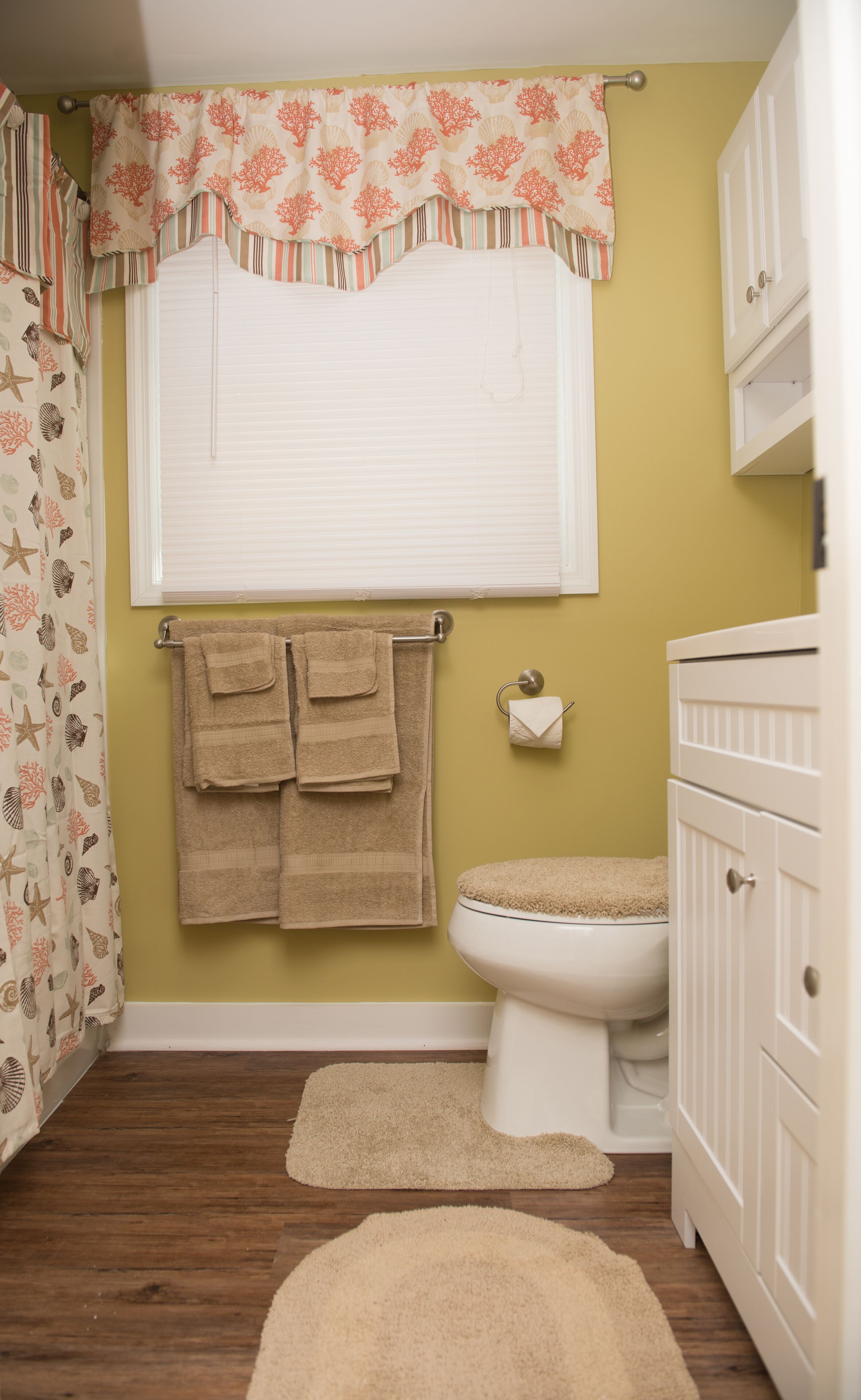 Une salle de bain comprenant des toilettes, un lavabo, un rideau de douche orné d’un motif de coquillages et un tapis beige.