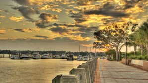 A waterfront marina with boats docked, a paved walkway, and trees along the edge.