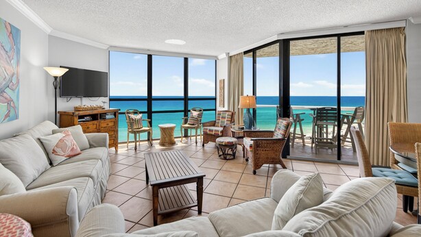 A spacious living room with a large TV, comfortable seating, and a balcony with a view of the ocean.