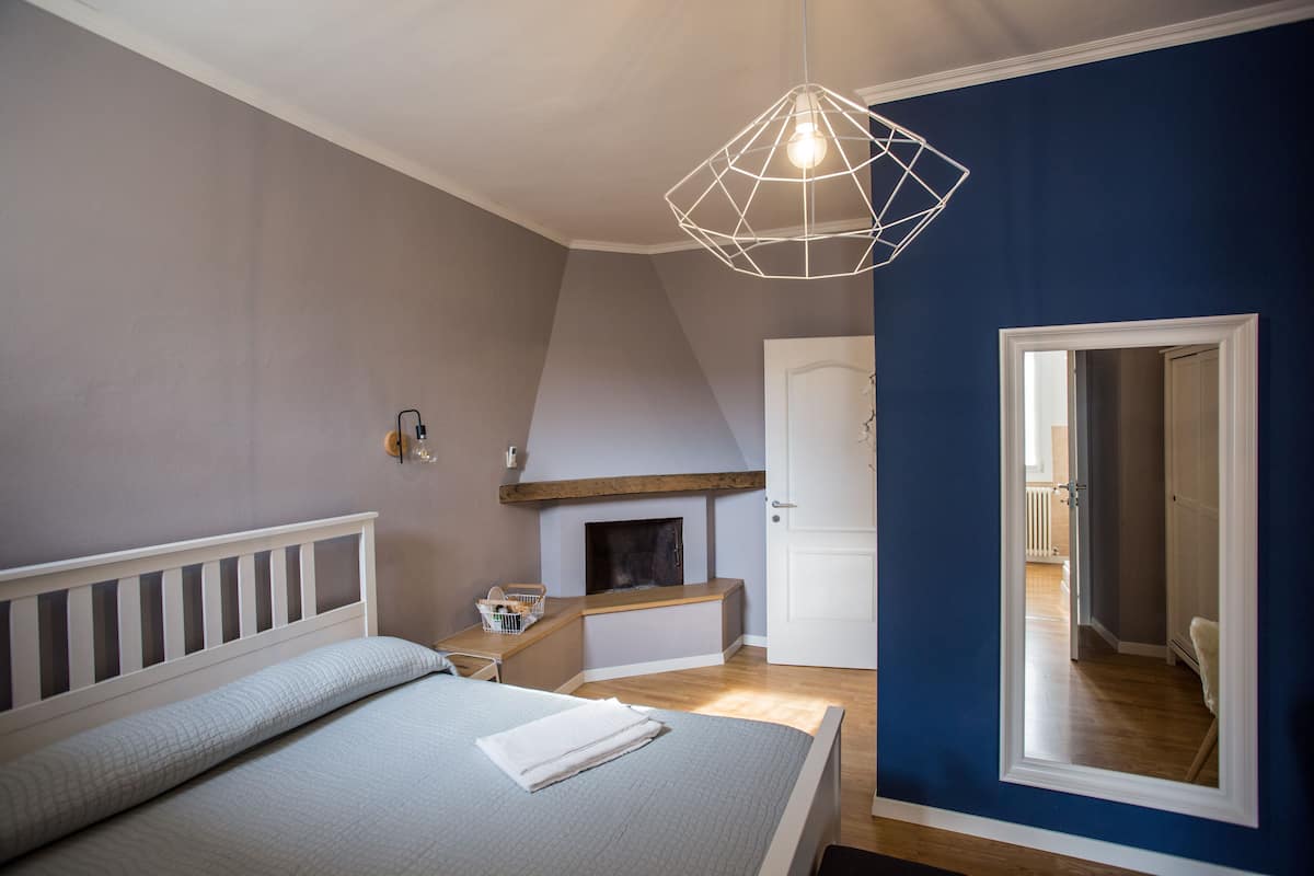 Une chambre à coucher comprenant un lit, un luminaire suspendu, une cheminée et une porte donnant sur une autre pièce.